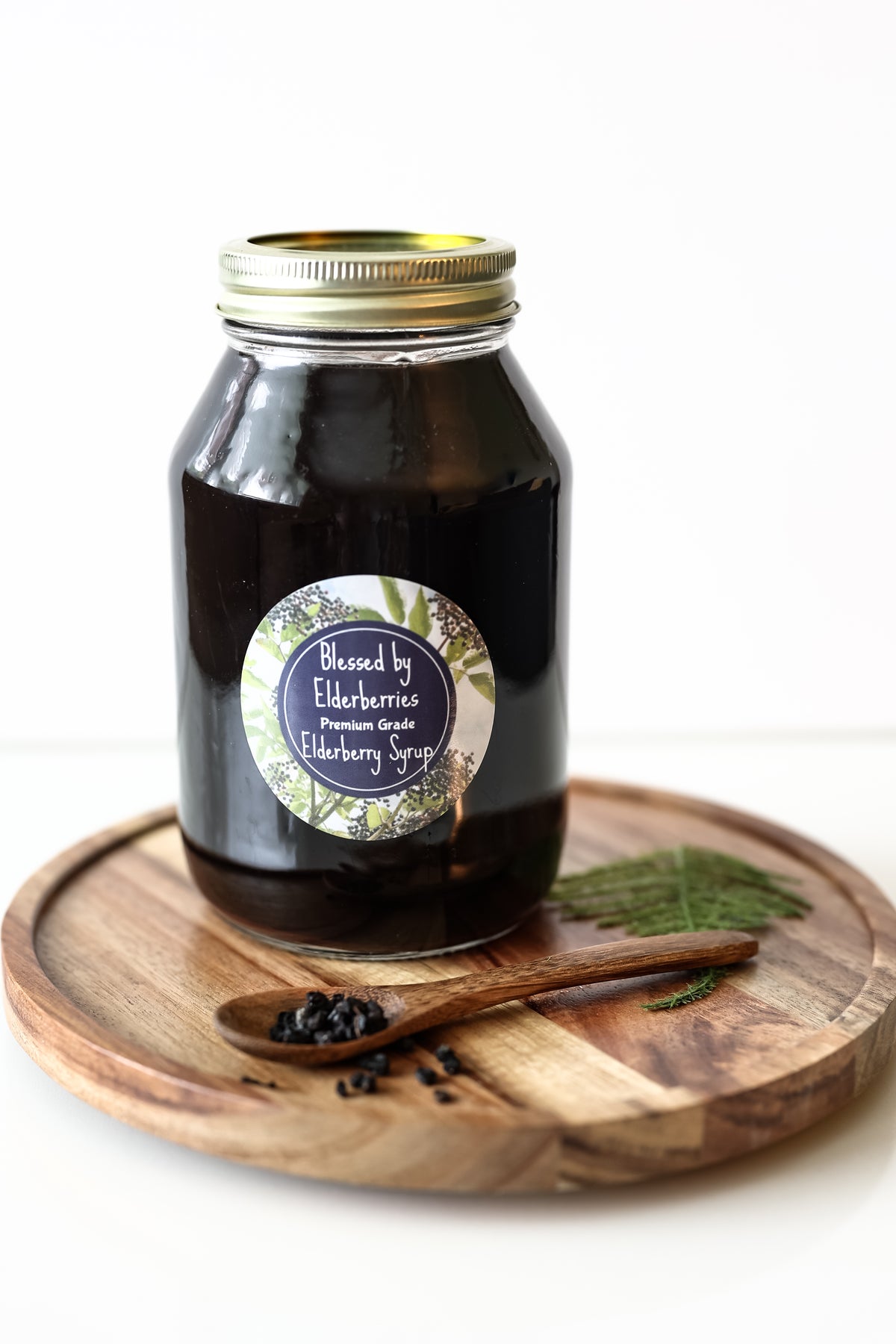 A 32oz glass jar of Blessed by Elderberries Premium Organic Elderberry Syrup with a gold metal lid and a round label, displayed on a wooden serving tray with a wooden spoon of dark syrup and fresh green leaves on a white background.