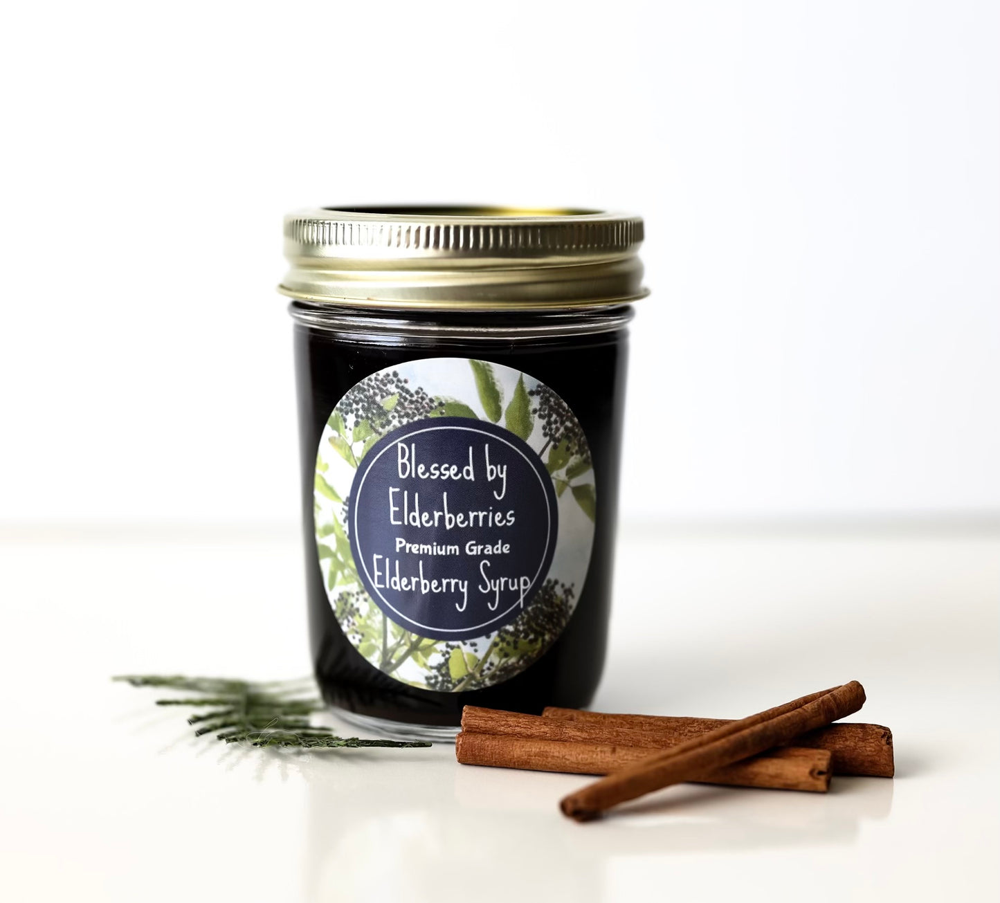 A glass jar of elderberry syrup with a gold metal lid and a circular label reading 'Blessed by Elderberries Premium Grade Elderberry Syrup,' styled on a white surface with cinnamon sticks and a sprig of greenery against a white background.